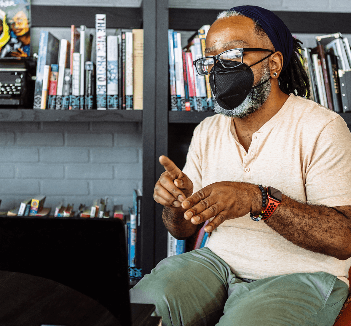 Portrait shot of a Deaf Black man wearing glasses and a KN-95 mask signing in a video chat via laptop. The man sits at a coffee table inside a community library packed with books and posters.