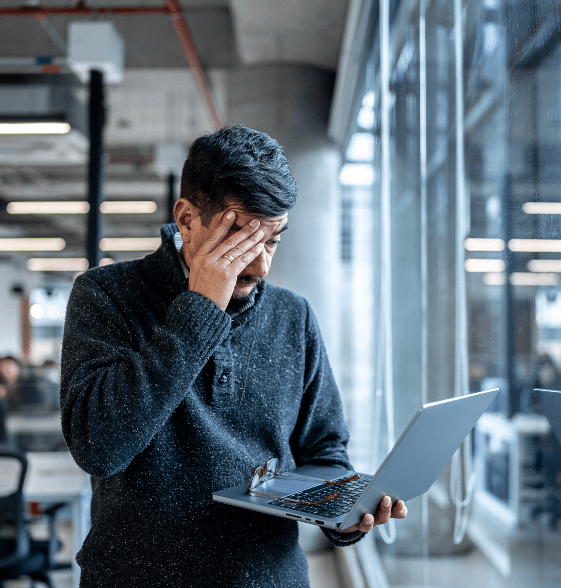 A man holding a laptop who is holding his hand to his face.