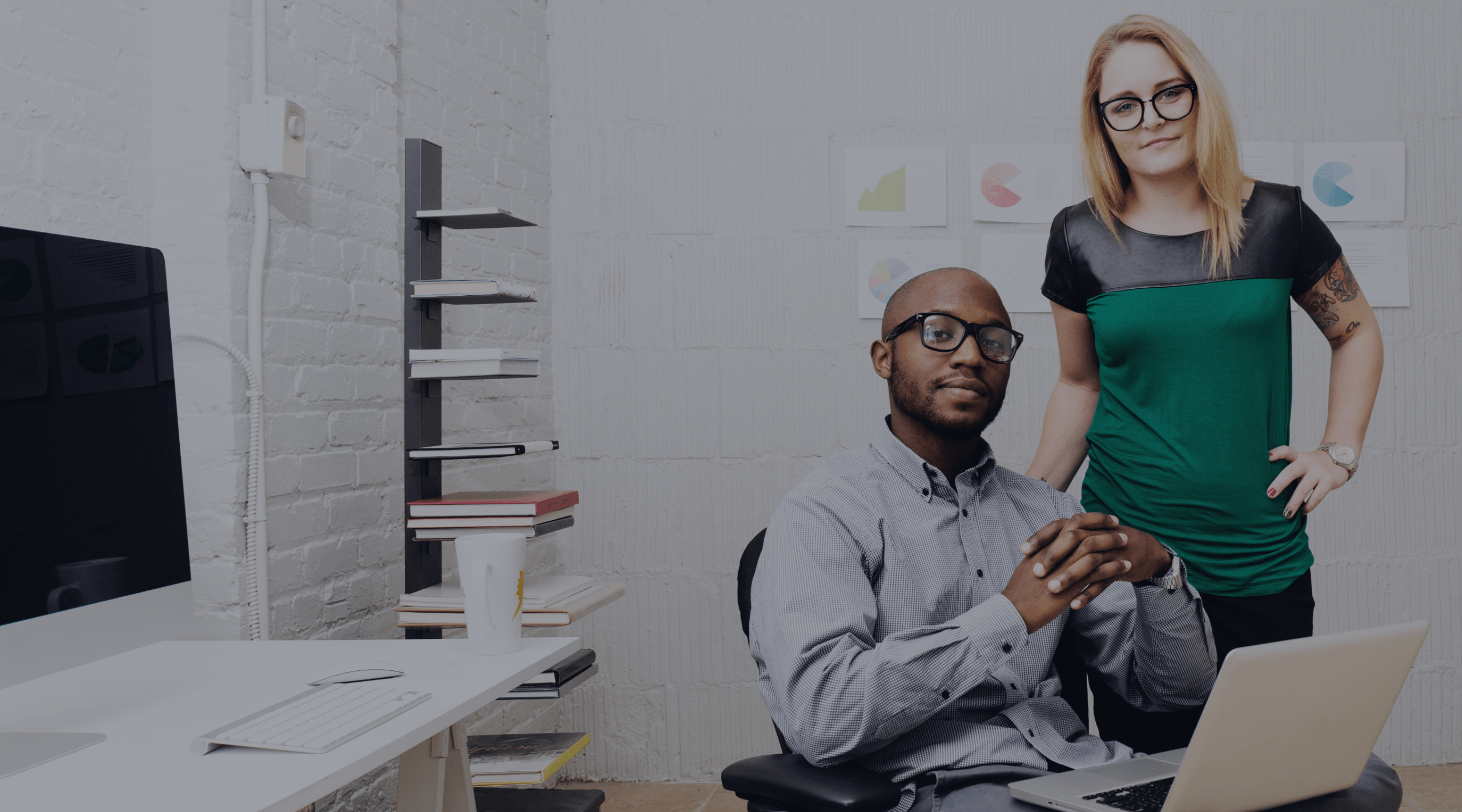 Man sitting in an office working on a laptop with women standing behind him.